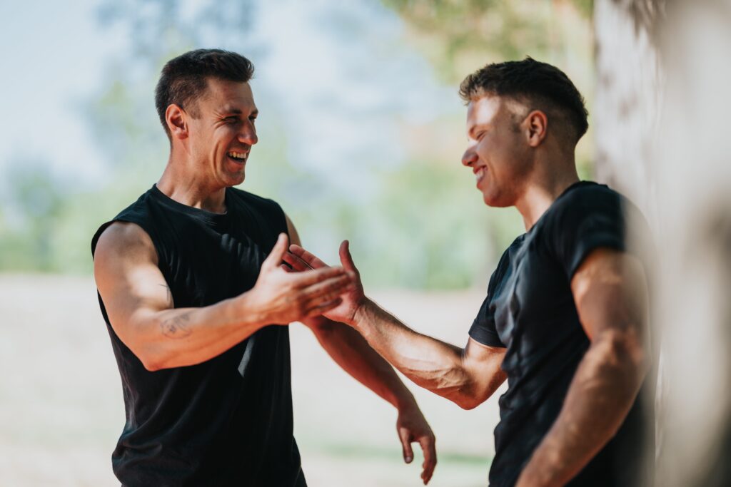 Two athletic men in black sleeveless shirts high-five outdoors after training, smiling and celebrating their workout together.
