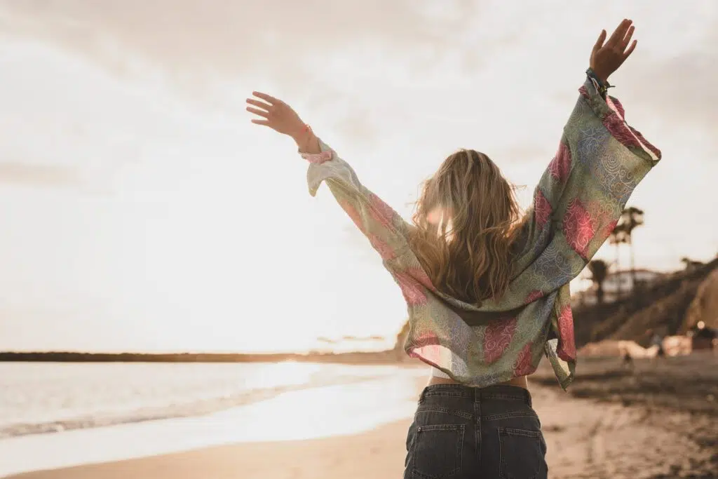 Woman standing on sandy beach at sunset facing the ocean with arms outstretched in joyful pose