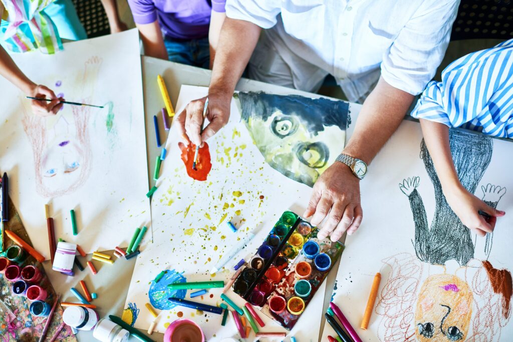 Top view of colorful drawings and paintings on a table with hands of students and teachers creating artwork in an art class