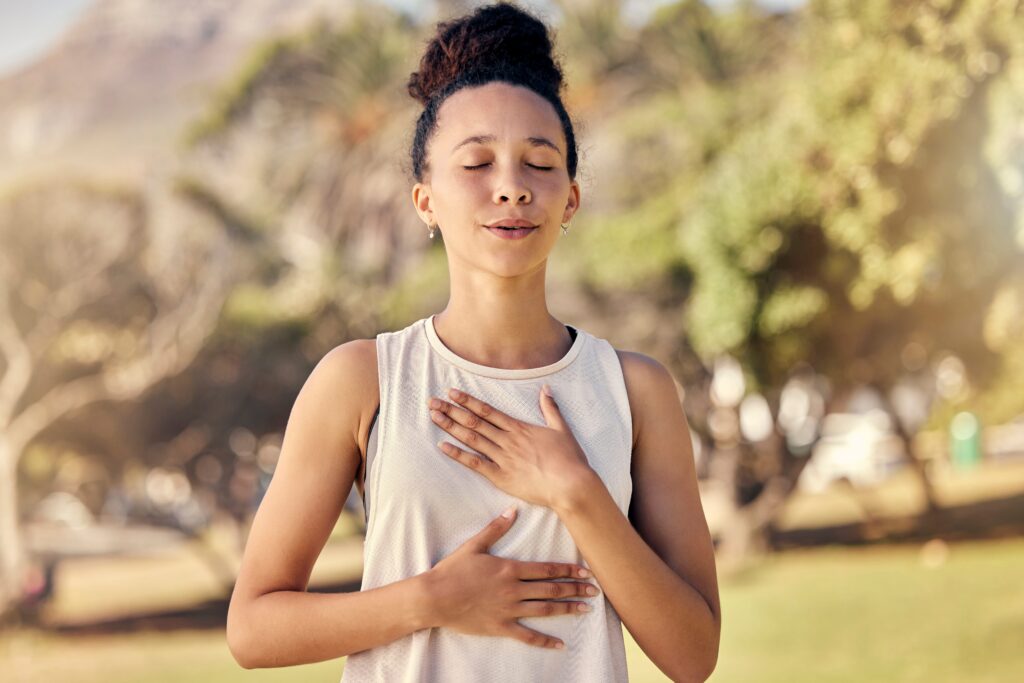 Woman in athletic wear practicing yoga breathing exercise outdoors with eyes closed and hands in prayer position at chest