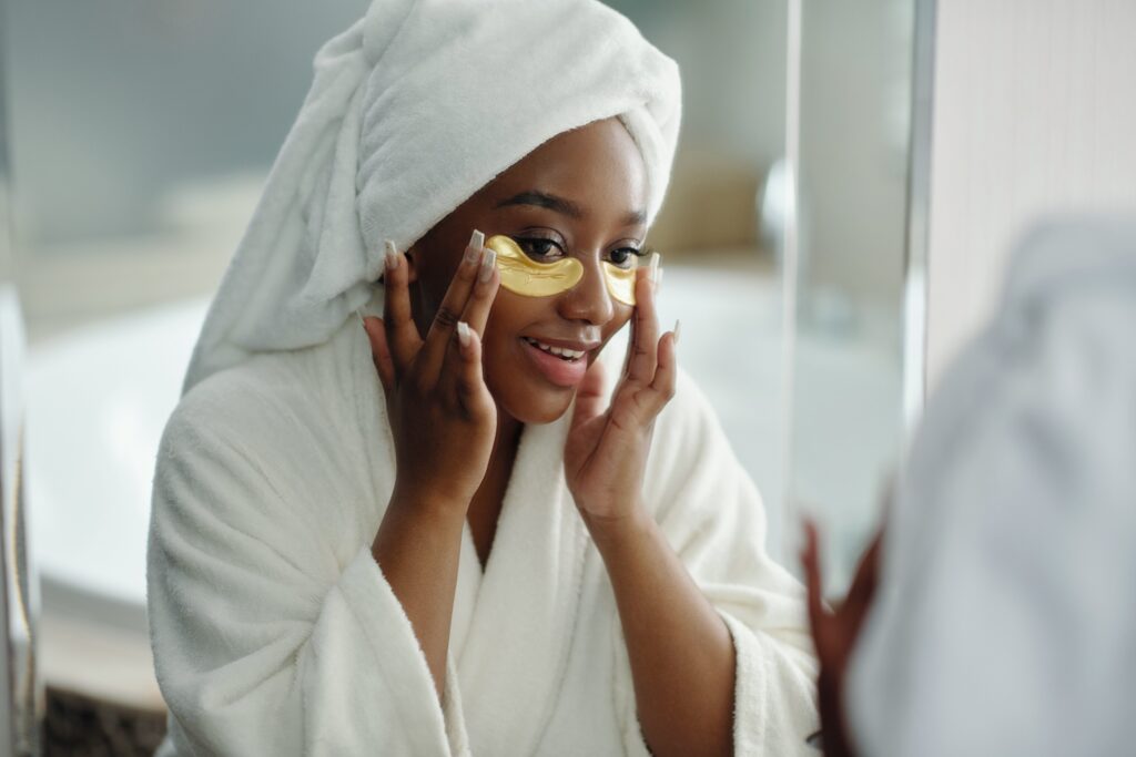 Woman with white towel wrapped on head applies hydrogel under-eye patches in bathroom, smiling during skincare routine