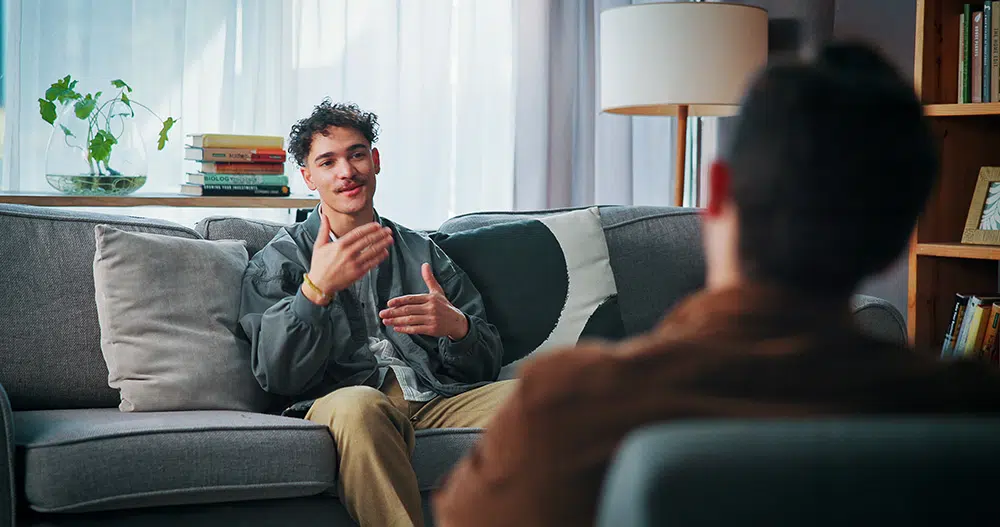 A man sits on a sofa speaking with a psychologist in a therapy office during a counseling session.