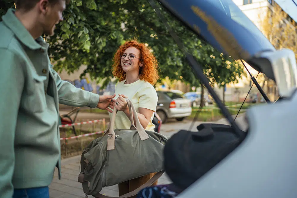 A man and woman place suitcases and bags into an open car trunk, preparing for a trip.