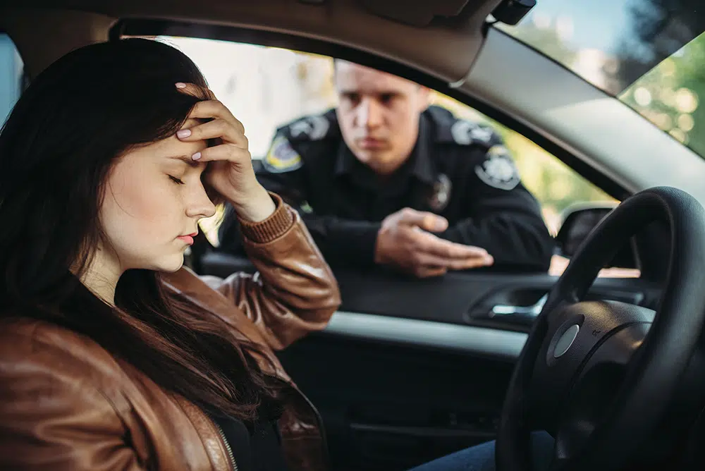 A male police officer leans toward a car window and speaks with a female driver during a traffic stop.