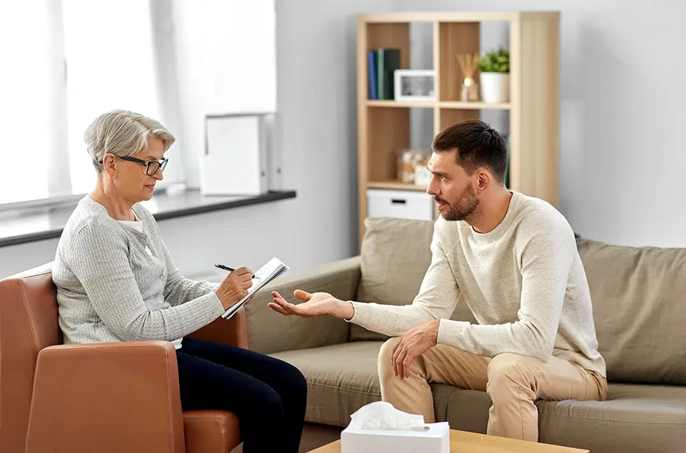 An older female psychologist takes notes while counseling a distressed young male patient during a therapy session.