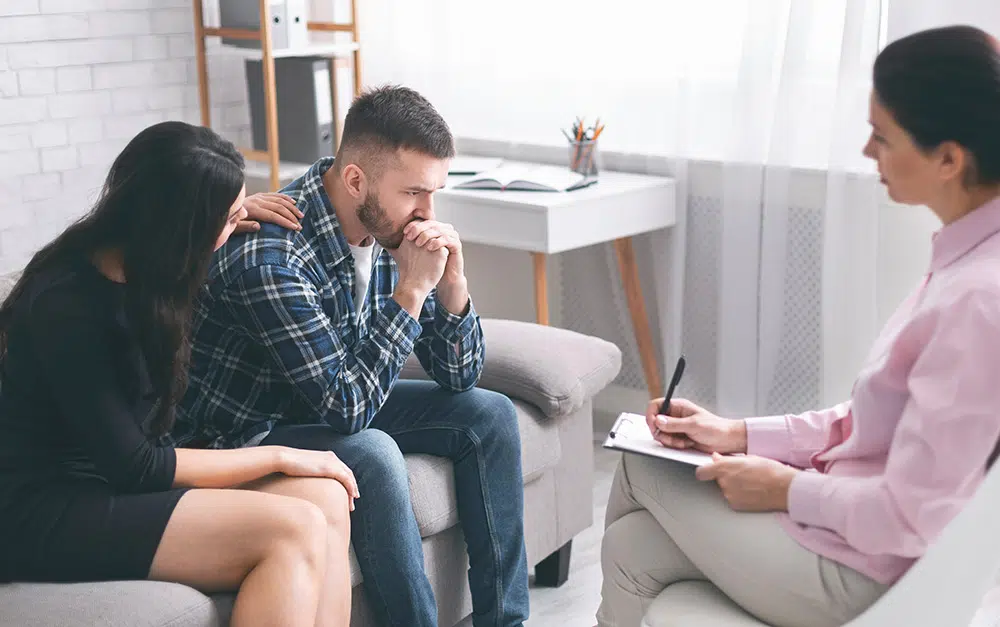 A woman places a comforting hand on a man's shoulder during a counseling session while a therapist observes with a clipboard.