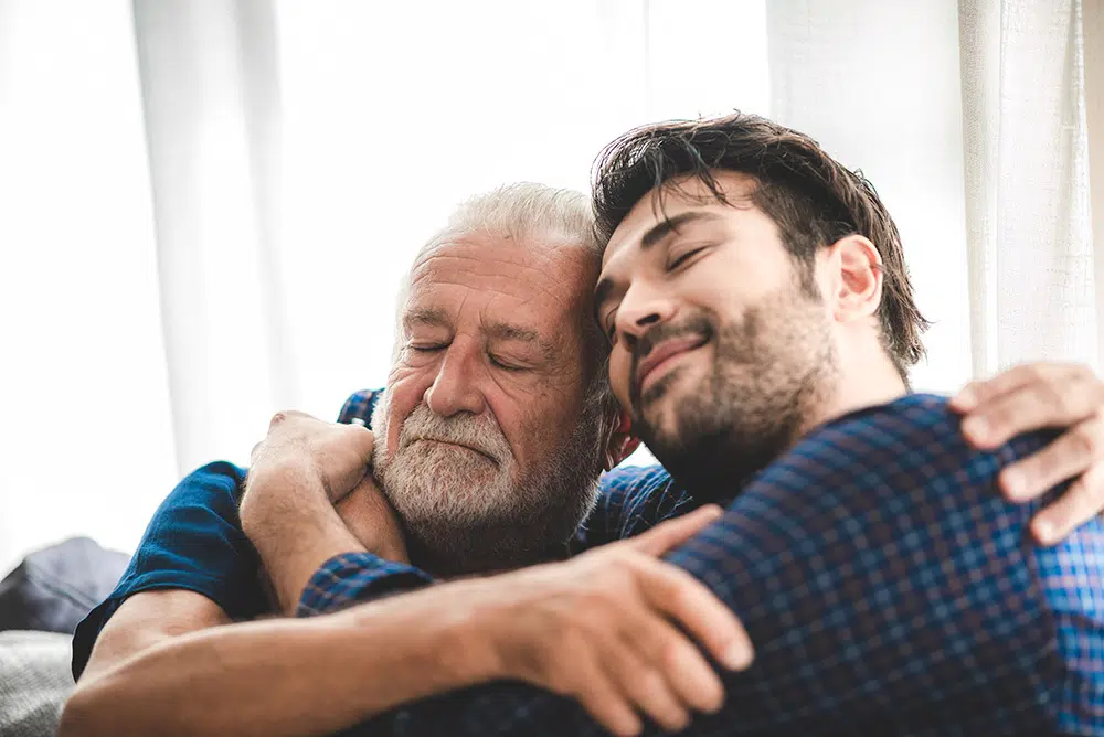 An adult son and senior father embrace warmly, while sitting together at home.