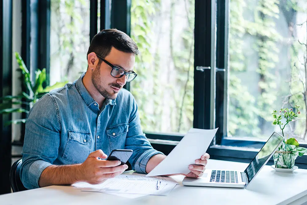 A young male works at laptop reviewing documents at an office desk.