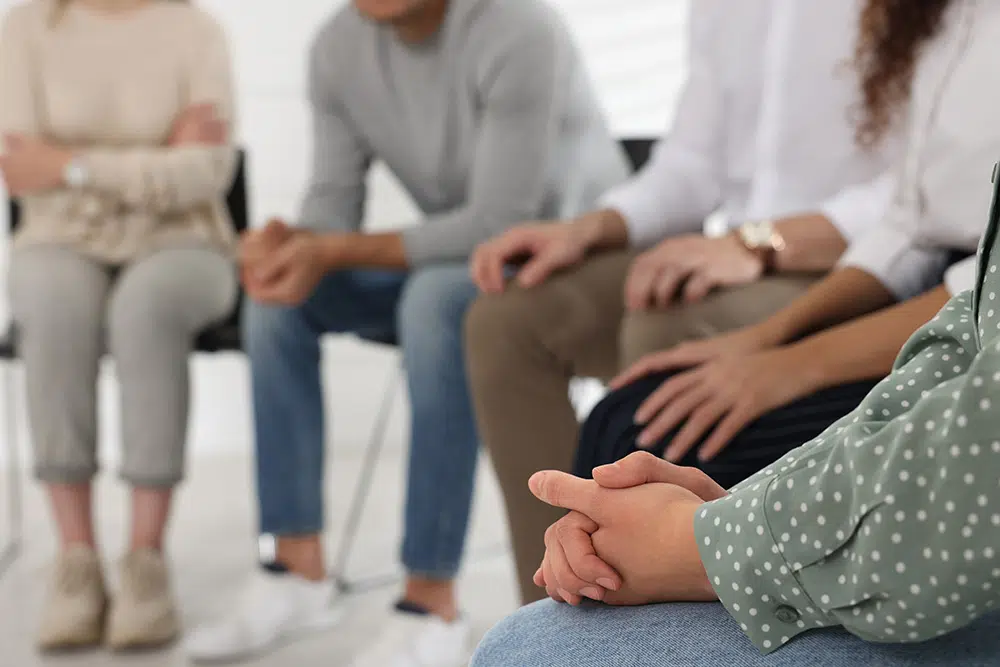 People sit in a circle during a group therapy session, closeup view of clasped hands and knees.
