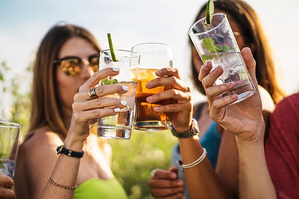 A group of young adults raise glasses in a toast outdoors on a sunny day, smiling and celebrating together.
