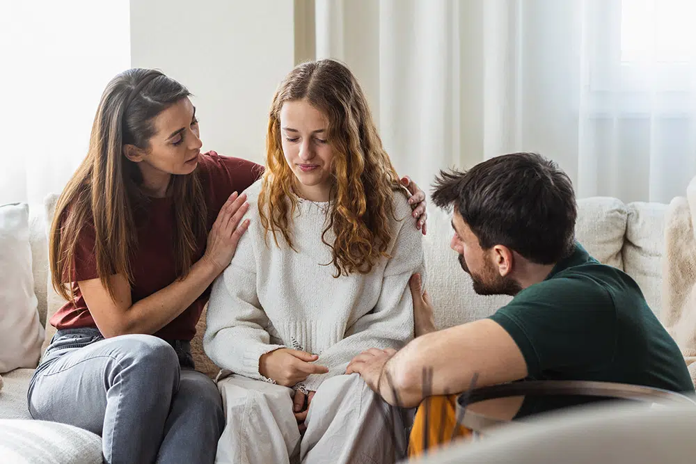 A teenage girl sits with her head down looking distressed while two adults, likely her parents, sit beside her offering comfort and support.