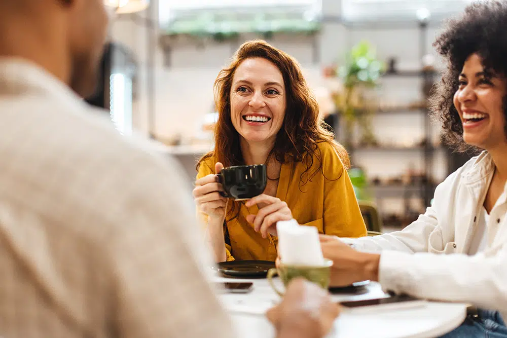 A group of young adults sit together at a table in a coffee shop, smiling and engaged in conversation.