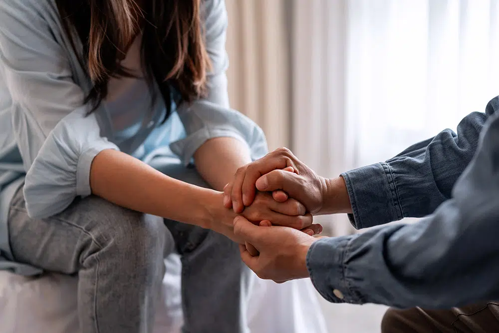 A man holds the hands of a distressed woman sitting on bed, offering emotional support in a home setting.