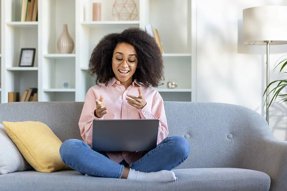 A woman smiles at a laptop screen while sitting on a gray sofa, engaging in a video call from home.