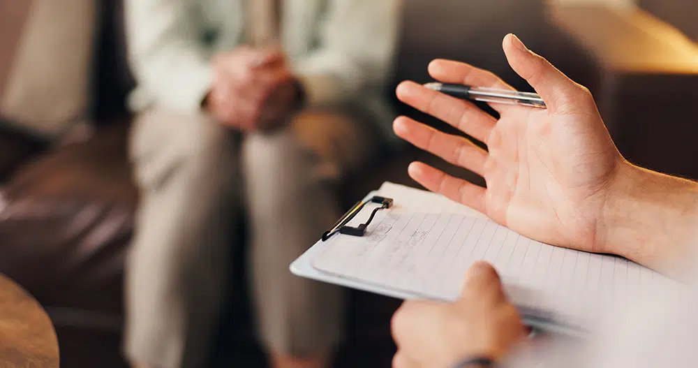 A therapist and patient's hands during a counseling session. The therapist holds a clipboard with notes while discussing mental health support.