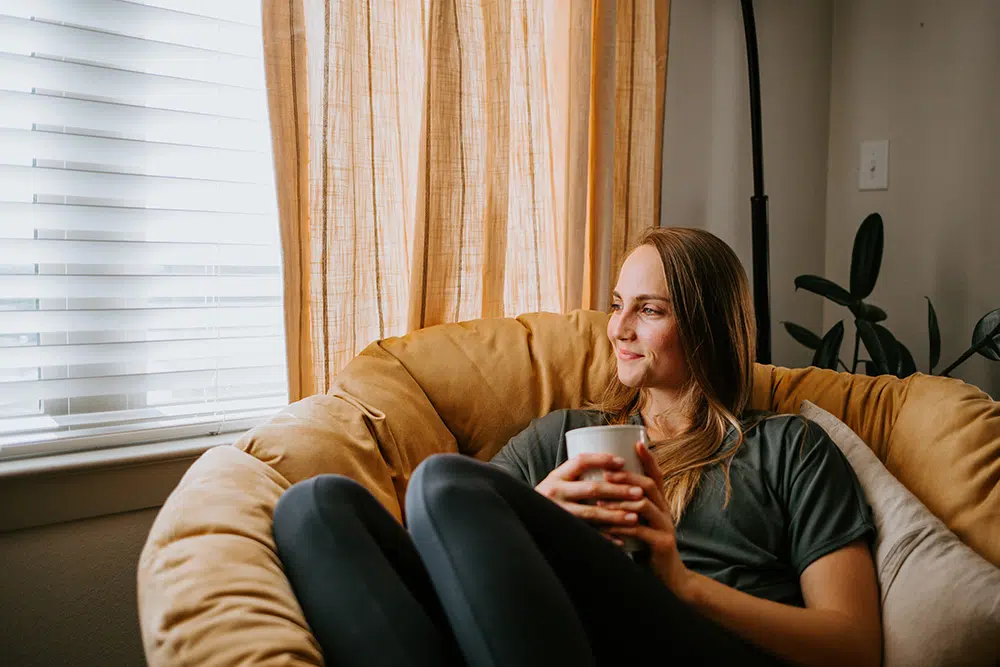 A woman in recovery sits on a sofa with a laptop and a mug. She is wrapped in blanket near a window with curtains.