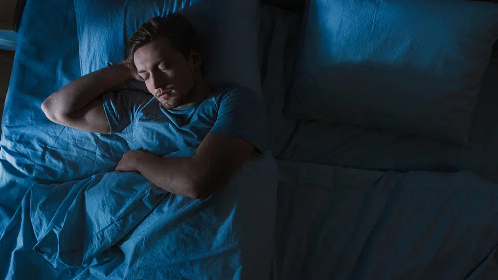 A young man in recovery sleeps in bed at night, viewed from above, with a blue lamppost light coming through window.