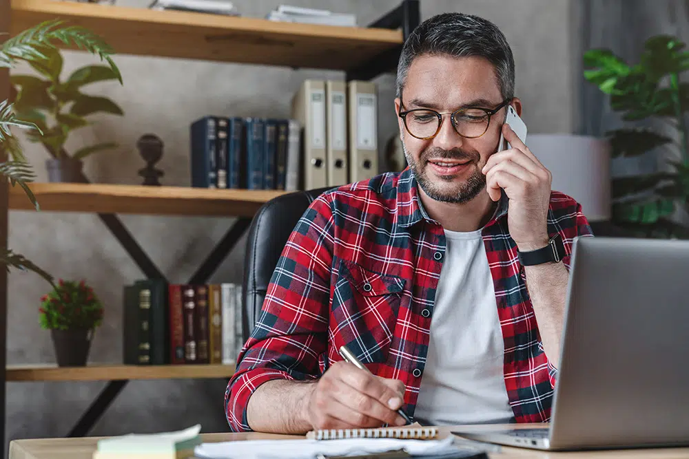 A smiling man writes notes while talking on phone and using a laptop at his home office desk.