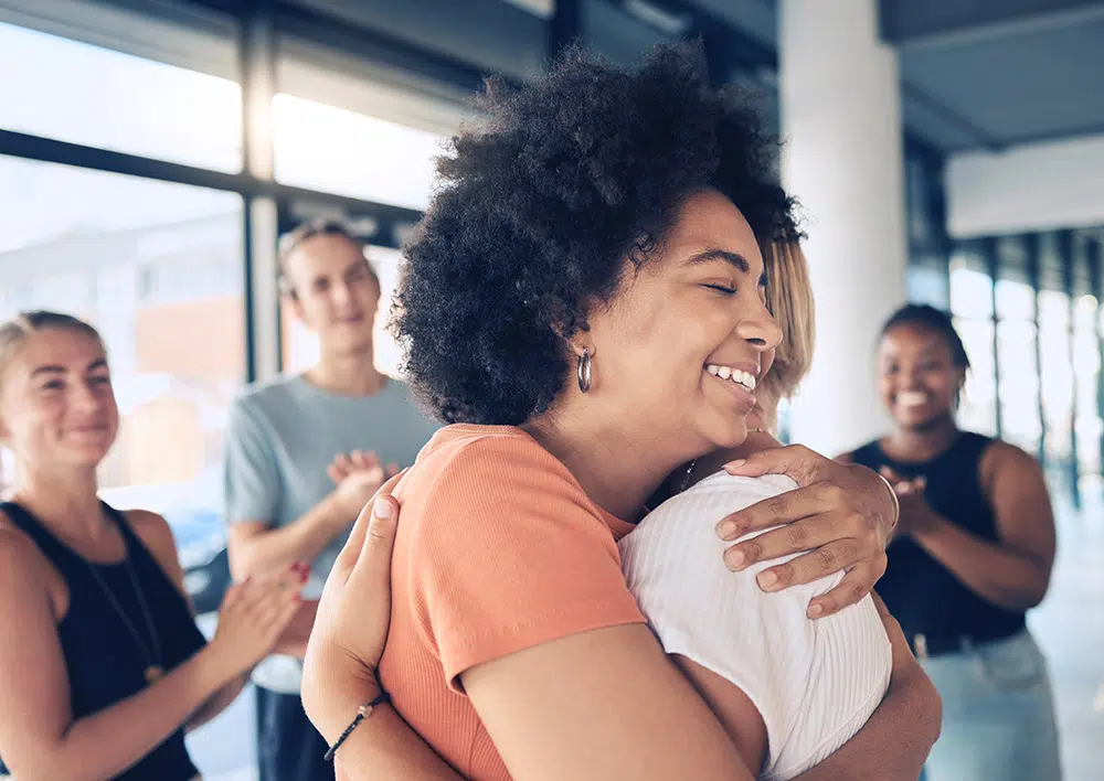 Two women embrace in foreground while group of people applaud in background during support group session.
