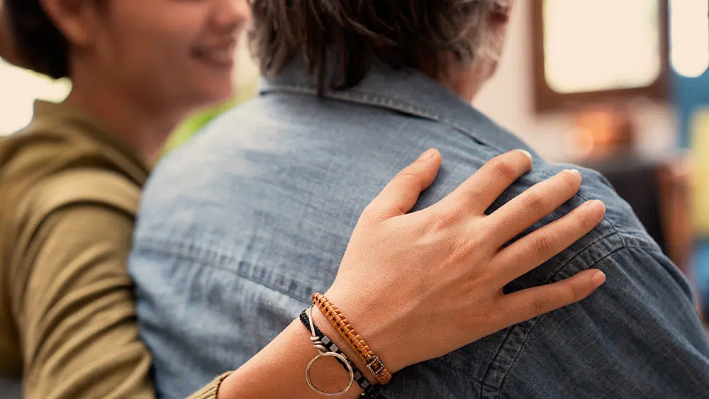 Close-up of the backs of two women with one hand over the other's shoulder in a bright, modern home interior.